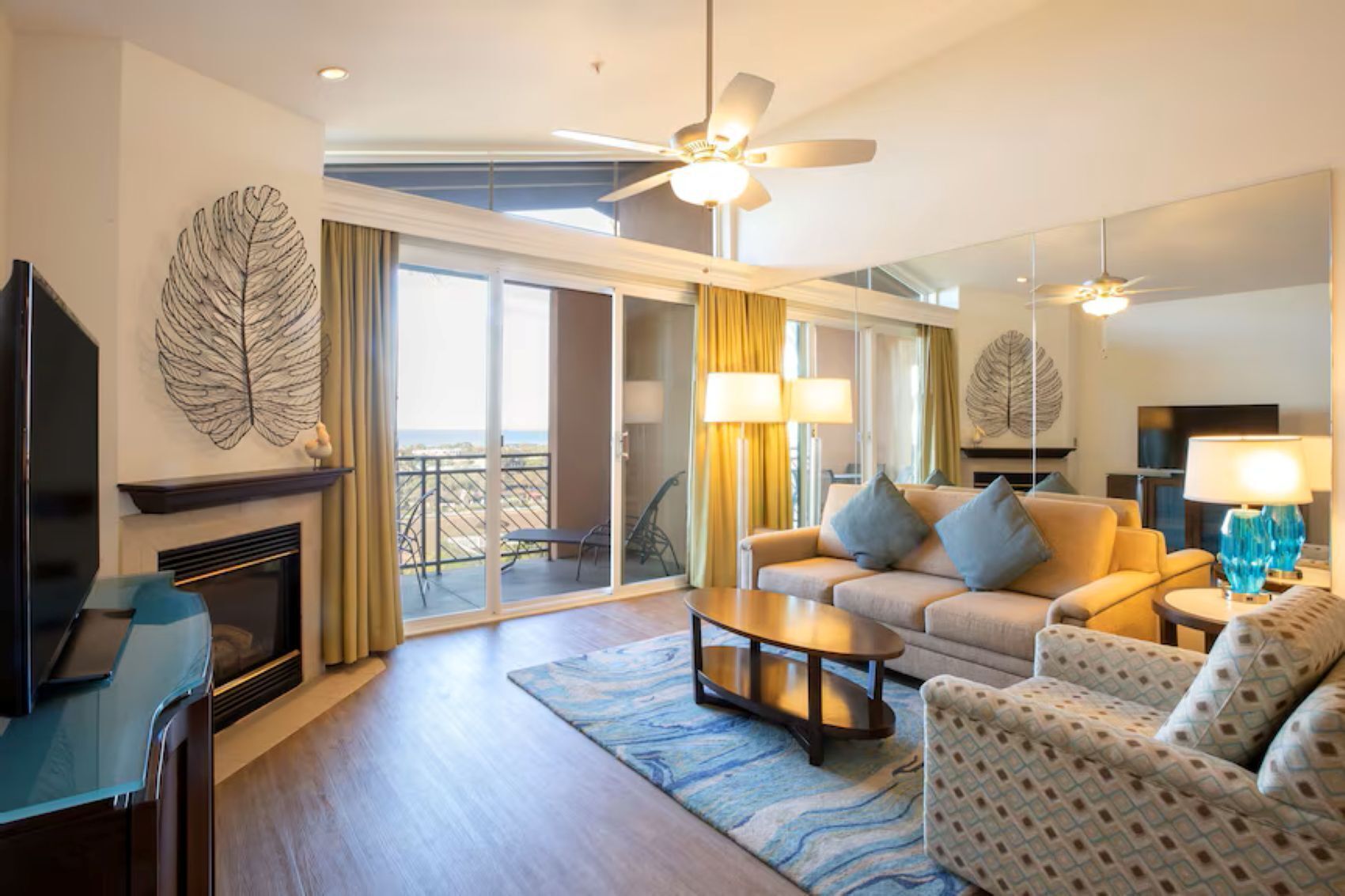 Quarto de hotel moderno com sofá, poltronas, TV, lareira, varanda com vista e ventiladores de teto.
