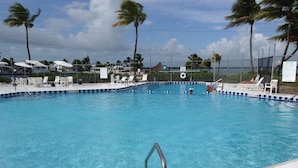 A swimming pool with a metal ladder, surrounded by white lounge chairs and umbrellas, with palm trees and a view of the ocean in the background.