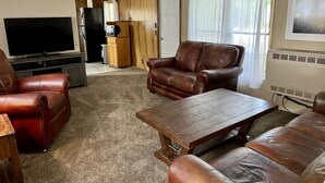 Una sala de estar con sofás de cuero, una mesa de centro de madera, un televisor de pantalla plana y una zona de cocina con refrigerador y microondas.
