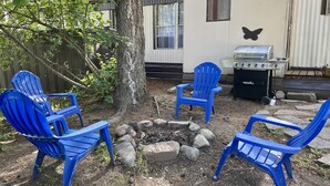 Patio trasero con fogón, sillas Adirondack azules, parrilla y casa rodante.