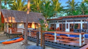 A beachside resort with thatched-roof structures, wooden deck seating, and a pool area.