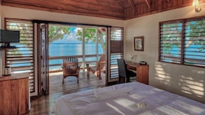 A bedroom with a large bed, a desk, a chair, and a TV. There is a sliding door leading to a balcony with a view of the ocean.