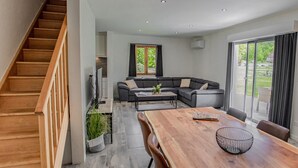 Una sala de estar moderna con una escalera de madera, un sofá modular, una mesa de comedor y un ventanal con vistas a zonas verdes.