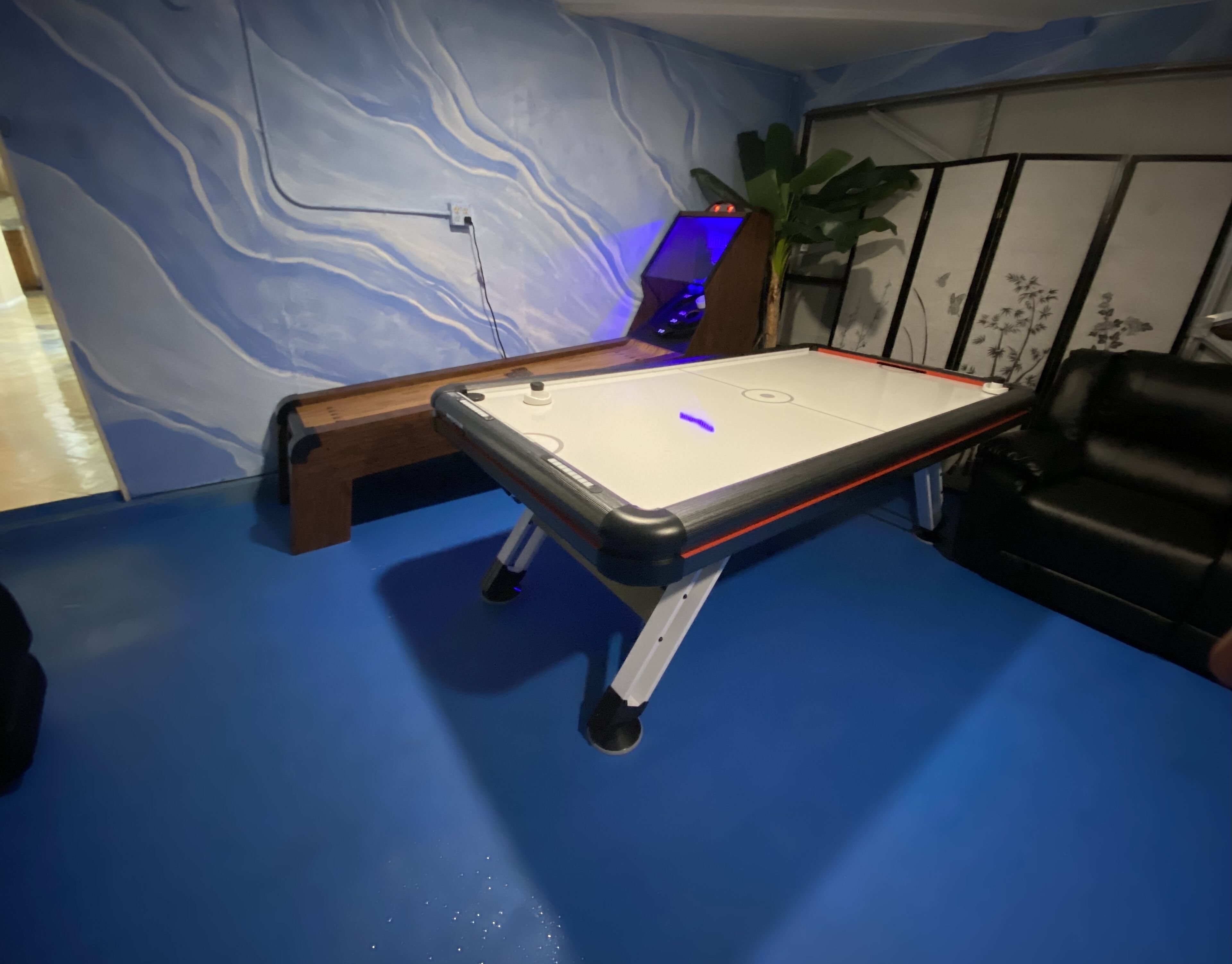 A room with a pool table, a blue wall with a wavy pattern, a plant, and a black leather chair.