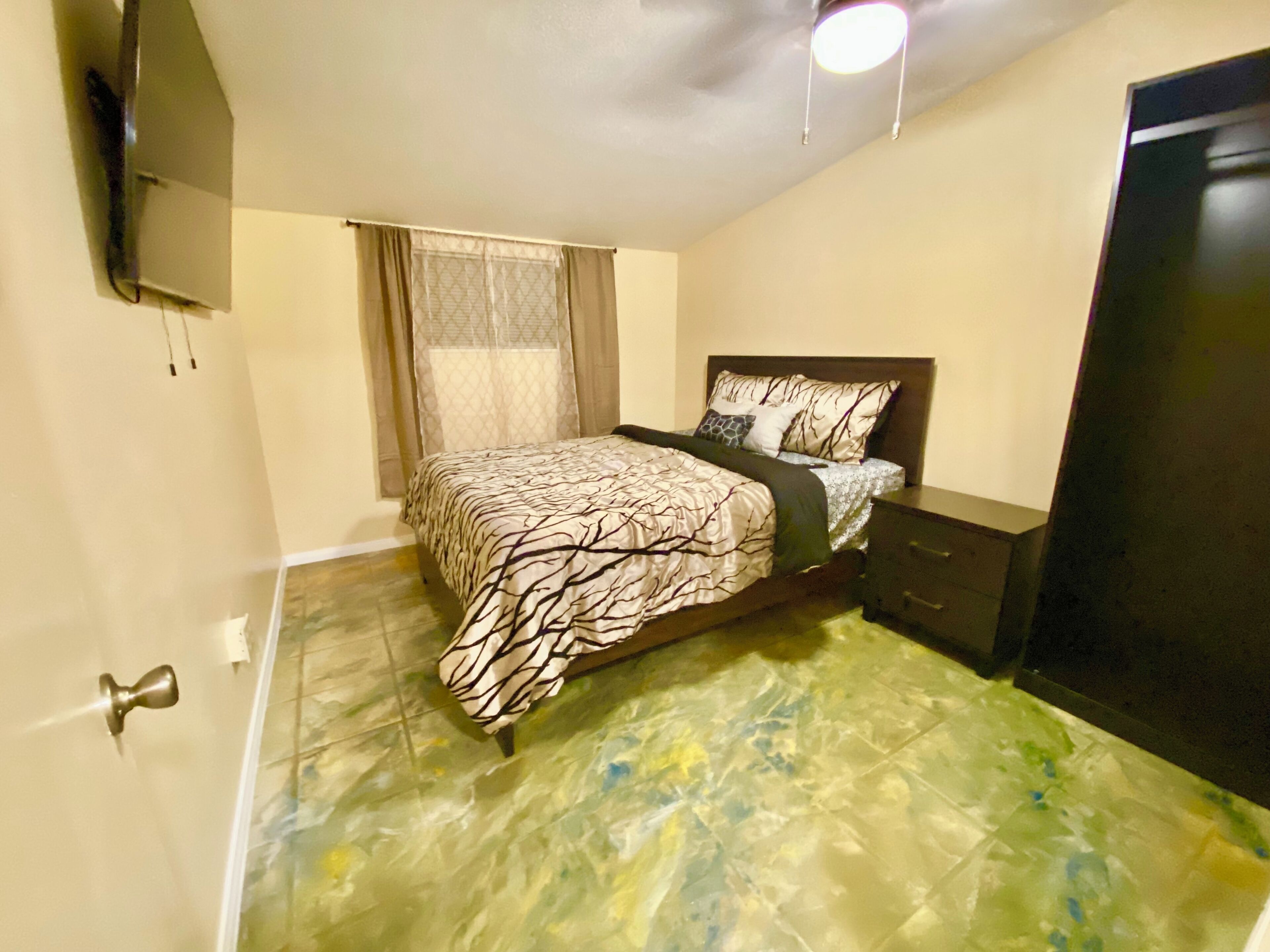 A bedroom with a bed, a nightstand, a dresser, a window with curtains, and a ceiling light.