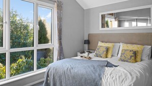 A bedroom with a large window, a bed with patterned pillows, a bedside table with a lamp, and a mirror above the bed. - Killington Lodge, Kentisbury Grange (Barnstaple)