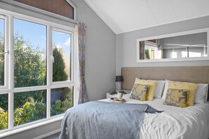 A bedroom with a large window, a bed with patterned pillows, a bedside table with a lamp, and a mirror above the bed.