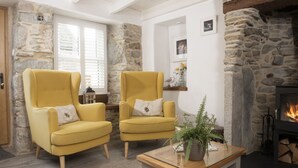 A cozy living room with two yellow armchairs, a small table with a plant, a stone fireplace, and a window with blinds. - Eastleigh Cottage - sleeps 4 guests  in 2 bedrooms (Marazion)