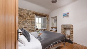 A bedroom with a stone wall, a fireplace, a bed with a gray blanket, and a window with shutters. - Eastleigh Cottage - sleeps 4 guests  in 2 bedrooms (Marazion)