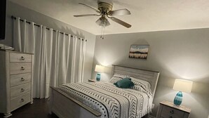 A bedroom with a bed, two bedside tables with lamps, a ceiling fan, a dresser, and a window with curtains. - Home in Groves (Groves)