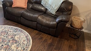 A brown leather sofa with two throw pillows, a woven basket on a small stool, and a patterned rug on a wooden floor. - Home in Groves (Groves)
