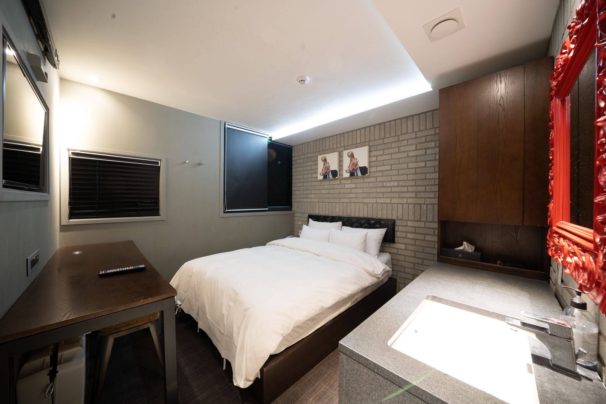A modern hotel room with a bed, a desk, a vanity with a mirror, and a brick-patterned wall.