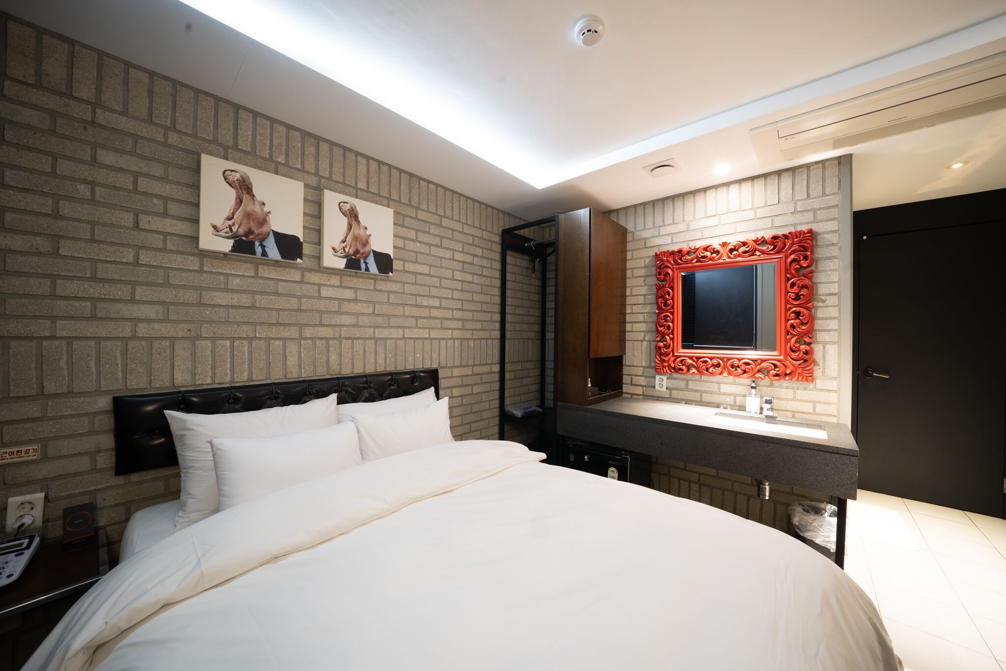Habitación de hotel con pared de ladrillo, cama con ropa de cama blanca, espejo grande con marco rojo y baño moderno con encimera oscura.