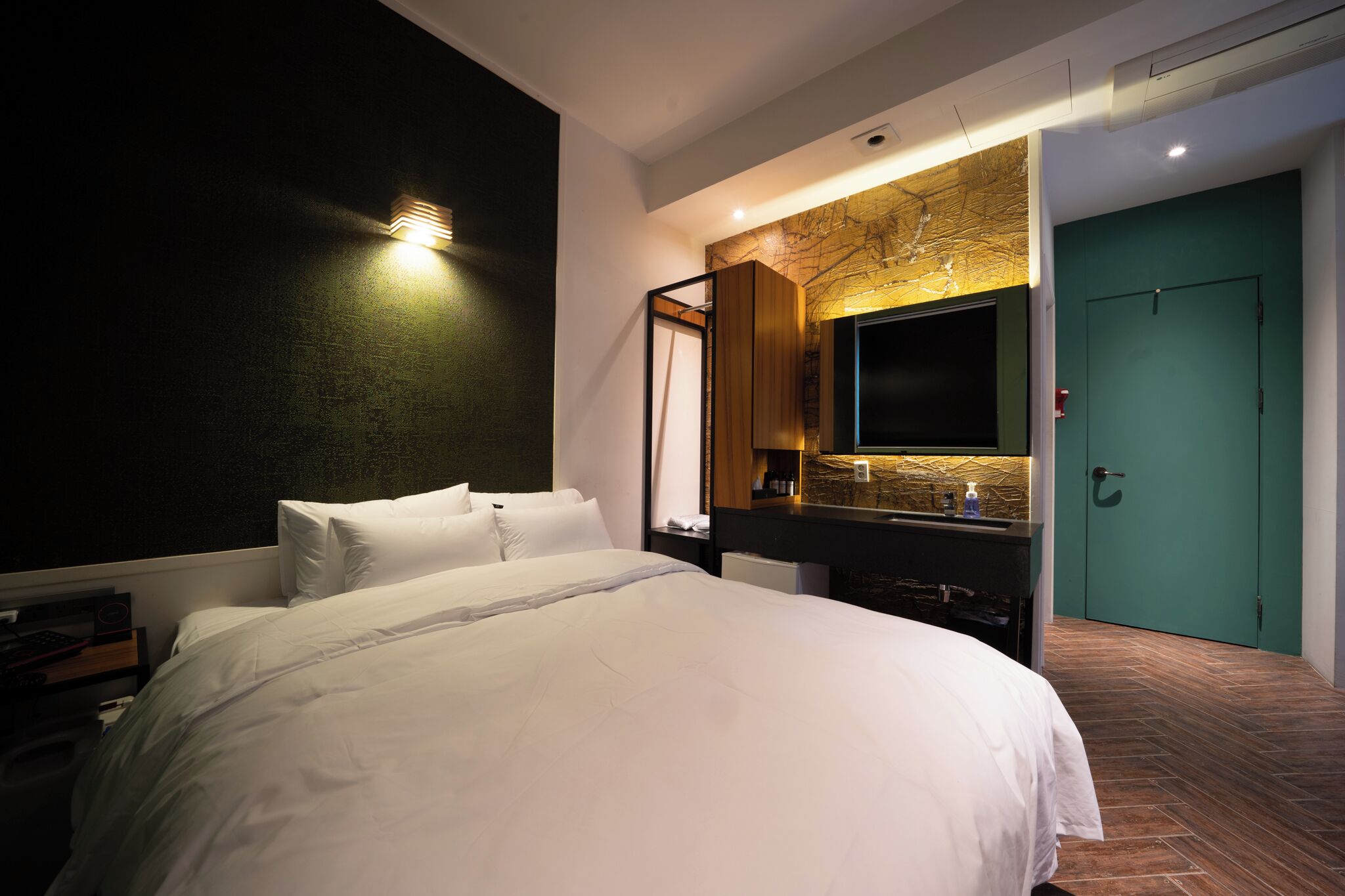 A modern hotel room with a large bed, a flat-screen TV, a vanity with a mirror, and a green door.
