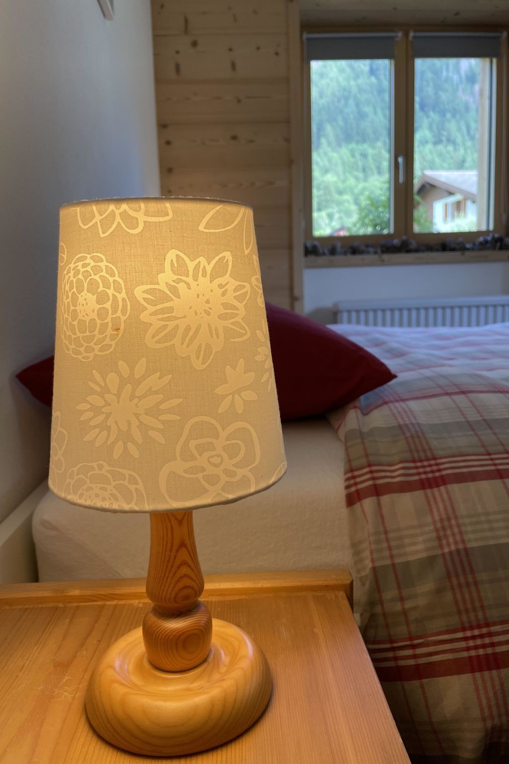 Una mesita de noche de madera con una lámpara con motivos florales. Una manta de cuadros cubre una cama con un cojín rojo.