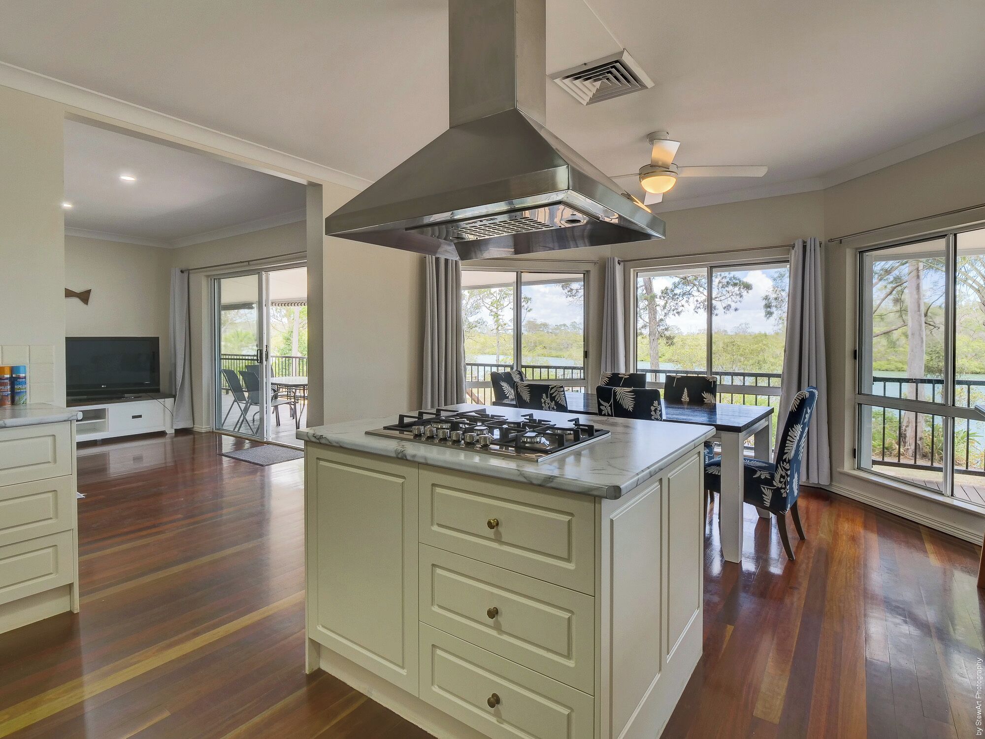 A spacious kitchen with a large island, a built-in stove, and a range hood. There is a dining area with a table and chairs, and large windows offering a view of the outdoors.