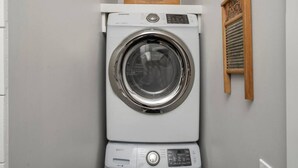 A front-loading Samsung washing machine in a laundry room with a wooden sign above it and a wooden hand dryer to the right.