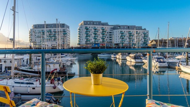 A balcony with a yellow table and a potted plant, overlooking a marina with boats and buildings in the background. - On the Water 4 By My Getaways (Brighton)