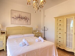 Een netjes opgemaakt bed met witte lakens en handdoeken, een kroonluchter, een stadsgezicht schilderij en een grote kledingkast.