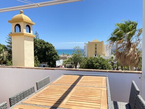 Een terras met een houten tafel en stoelen, met uitzicht op een kustgebied met gebouwen en palmbomen.