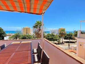 Een balkon met uitzicht op zee, een houten tafel, rieten stoelen en een gestreepte luifel.