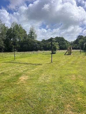 A grassy field with playground equipment, including swings and a slide, surrounded by trees and a clear sky.