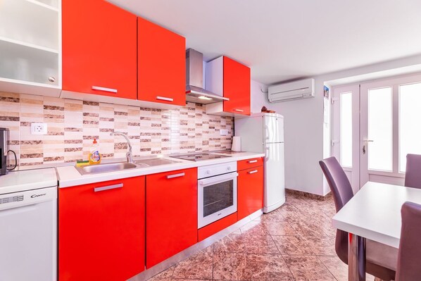 A modern kitchen with red cabinets, a white countertop, and a built-in oven. There is a dining area with a table and chairs.
