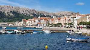 A coastal town with buildings along the waterfront, boats docked, and a mountainous backdrop. - First row to the sea (Baska)