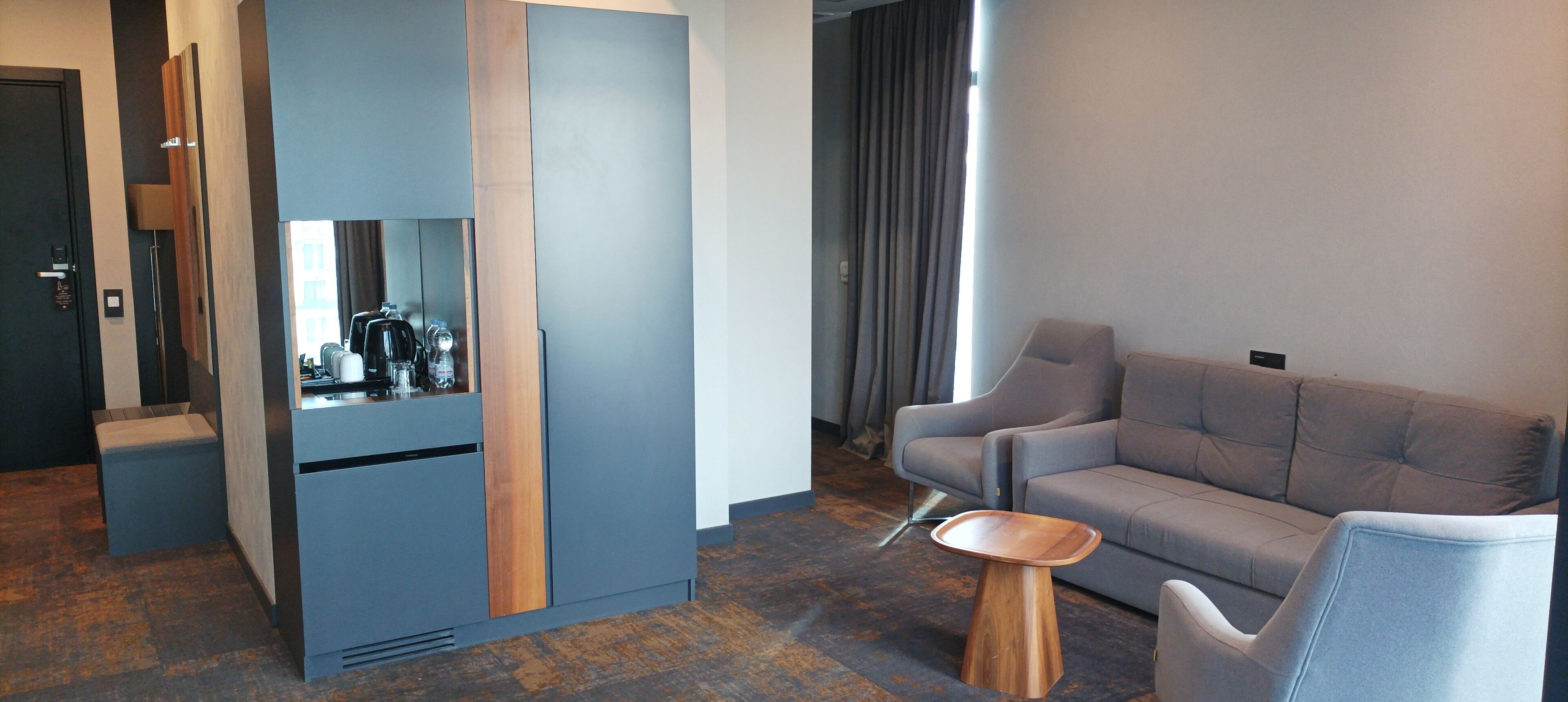 A modern hotel room with a grey sofa, a wooden side table, and a built-in coffee machine.