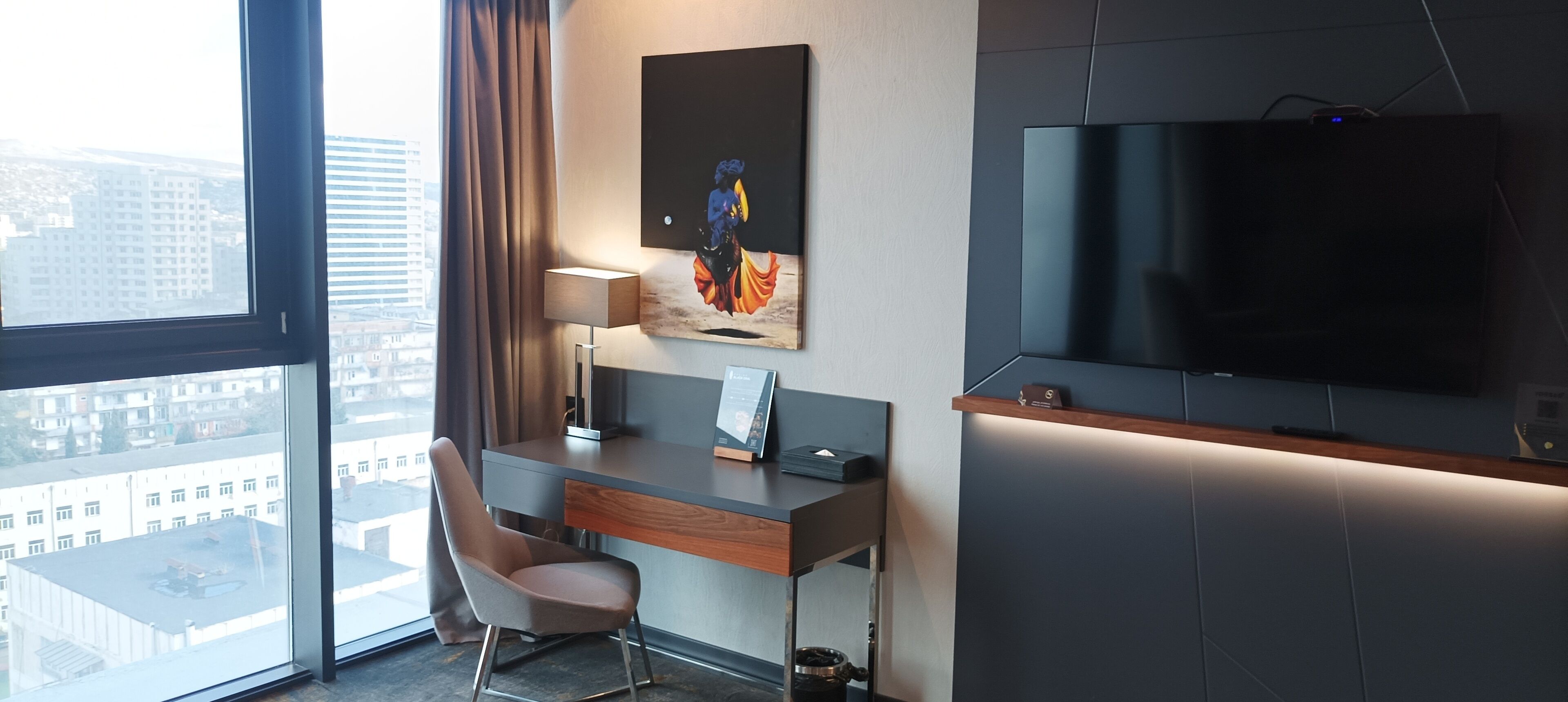 Habitación de hotel moderna con un gran ventanal, un escritorio con silla, un televisor de pantalla plana y un cuadro en la pared.