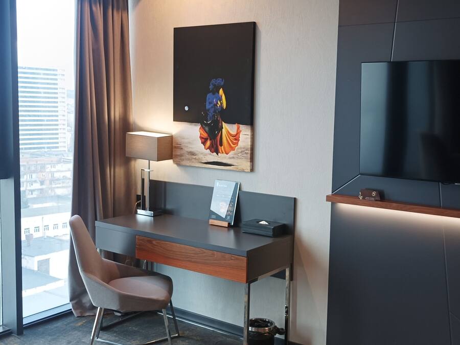 A modern hotel room with a large window, a desk with a chair, a flat-screen TV, and a painting on the wall.