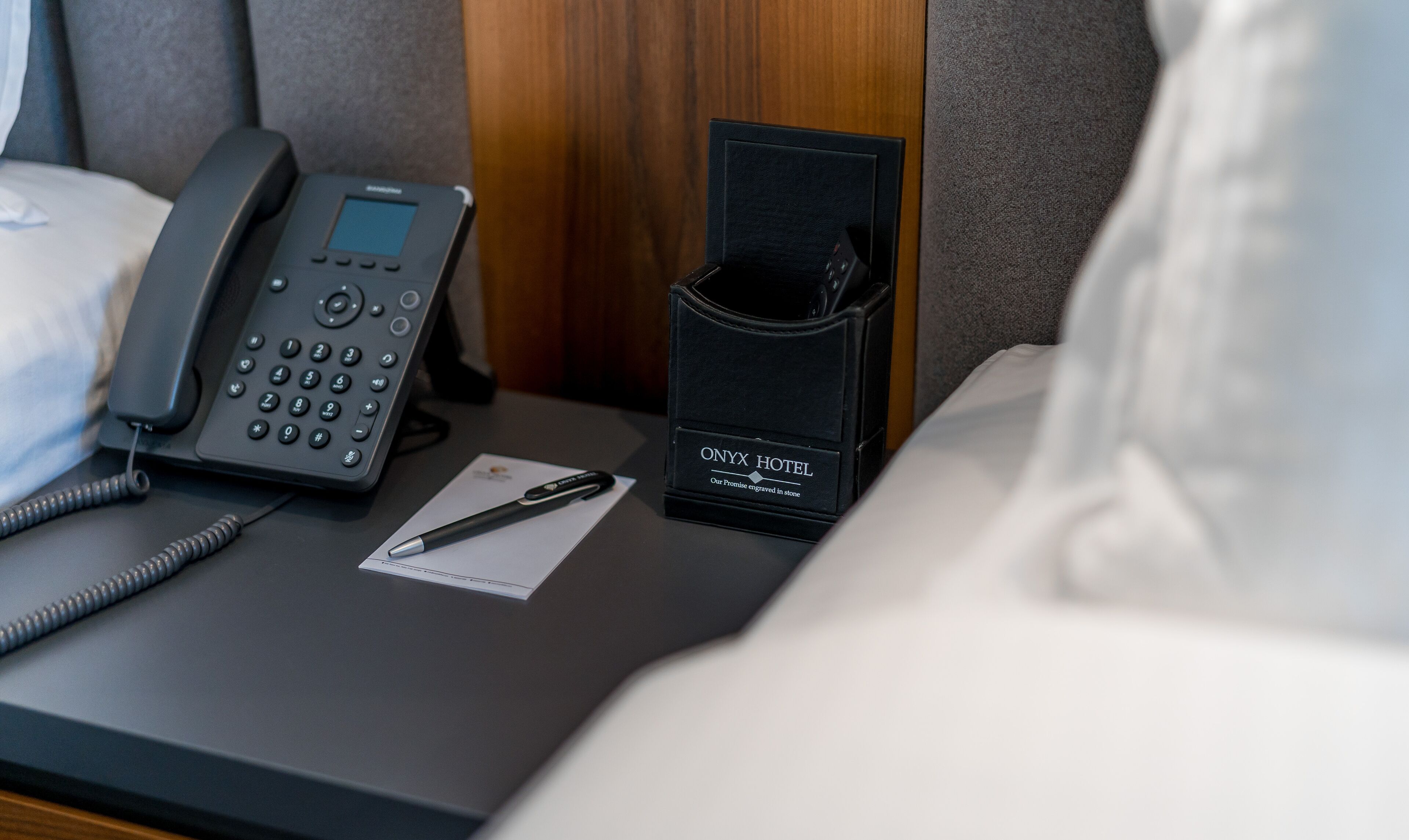 A hotel room with a bedside phone, a pen, and a black wallet with the hotel name on it.