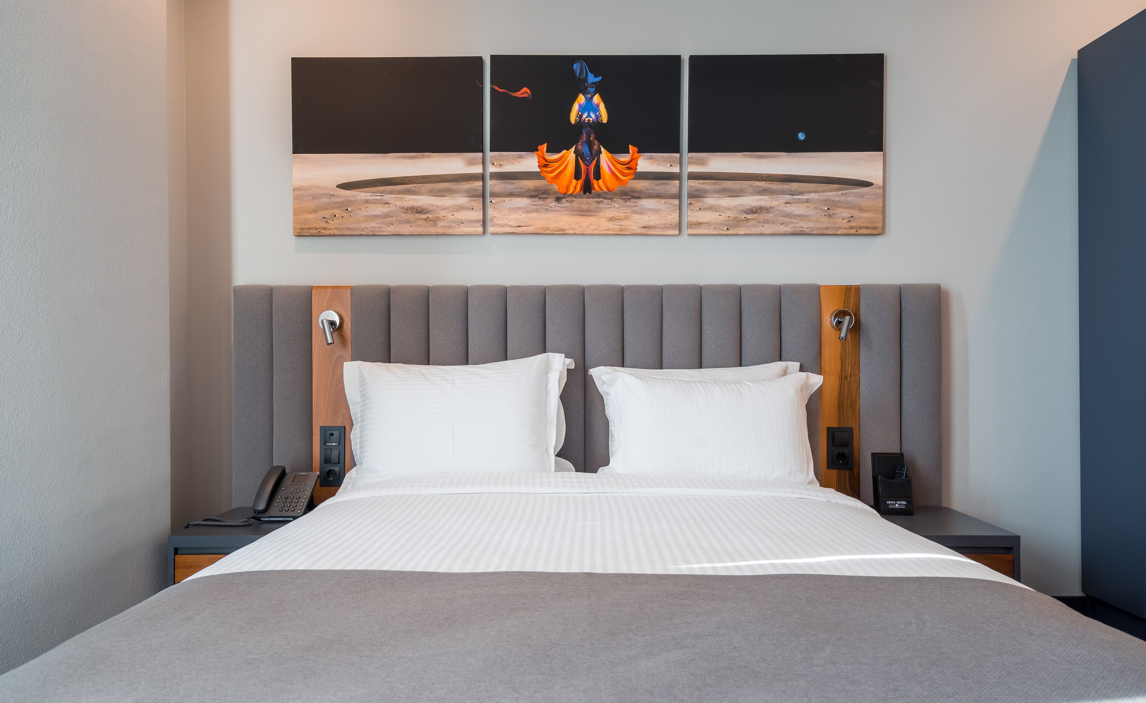 A hotel room with a large bed, a grey upholstered headboard, a bedside table with a phone, and a wall-mounted artwork depicting a figure on a planet.