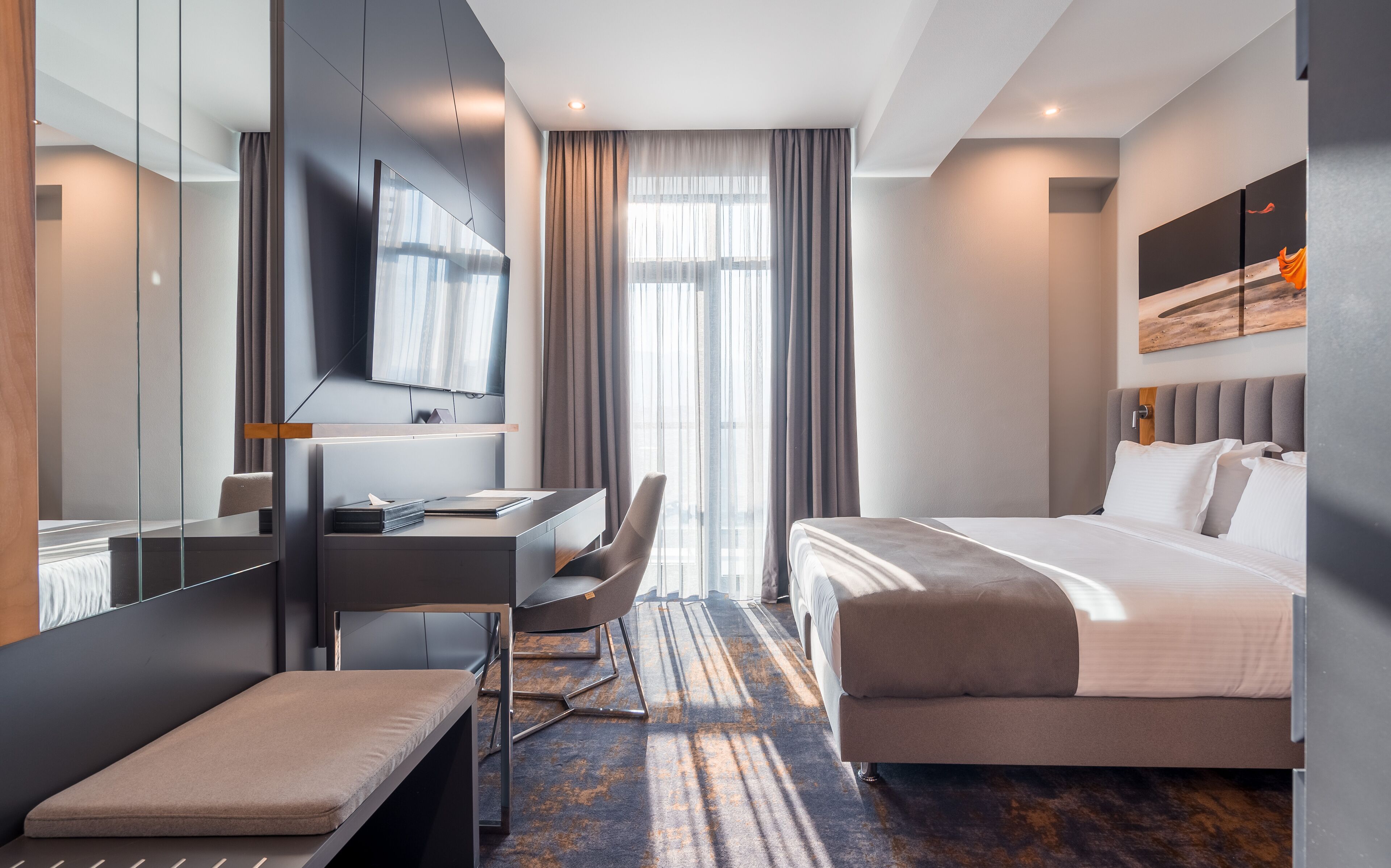 A modern hotel room with a bed, desk, chair, and a large mirror.