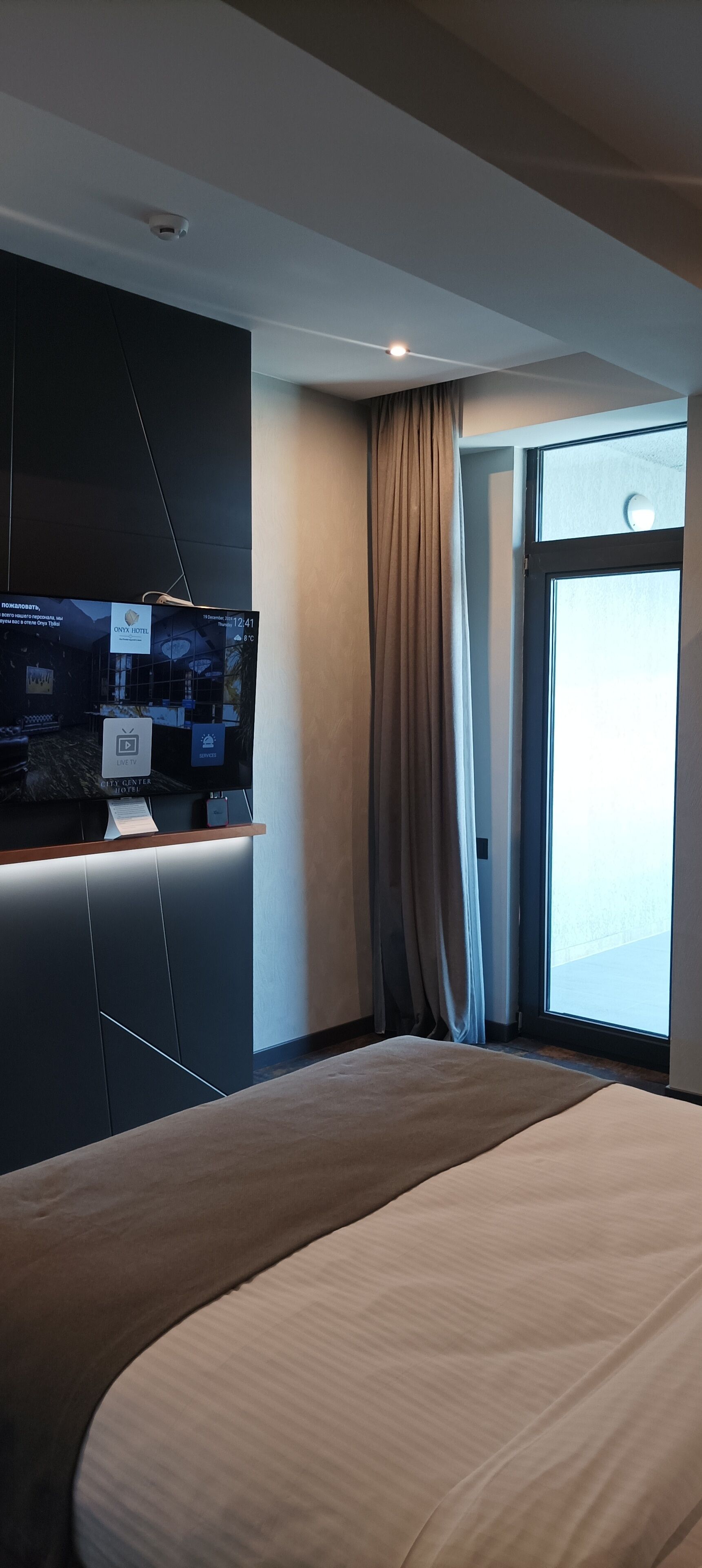 Una habitación de hotel moderna con una cama grande, un televisor de pantalla plana montado en la pared y un ventanal con cortinas.