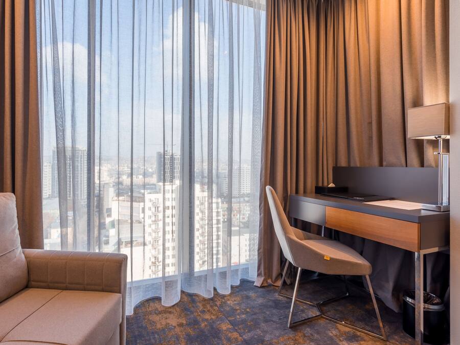 A hotel room with a large window, a sofa, a desk with a chair, and a TV.