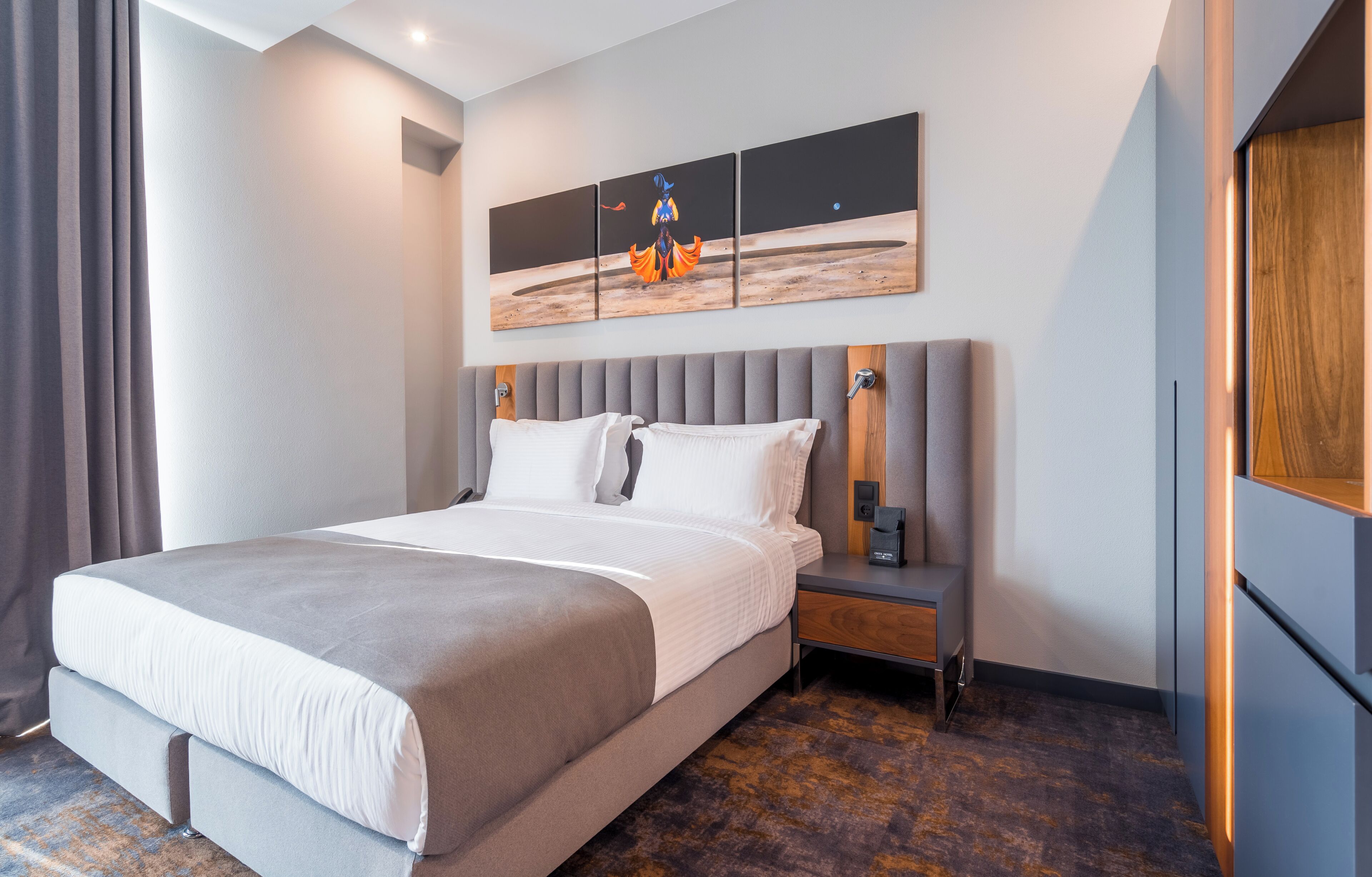 A modern hotel room with a large bed, a nightstand, a wall-mounted artwork, and a wooden panel on the right wall.