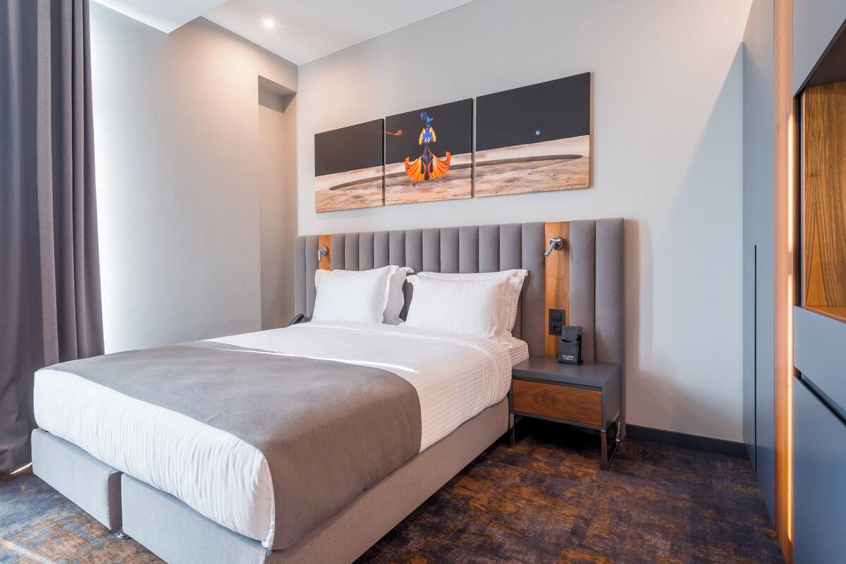 Moderni hotellihuone, jossa on suuri sänky, yöpöytä, seinälle kiinnitetty taideteos ja oikean seinän puupanelointi.