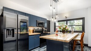 A modern kitchen with a stainless steel refrigerator, a microwave, and a coffee maker. There is a wooden island with a sink and bar stools. - Casa Del Exotique | Walk Everywhere | AC | Beach (Traverse City)