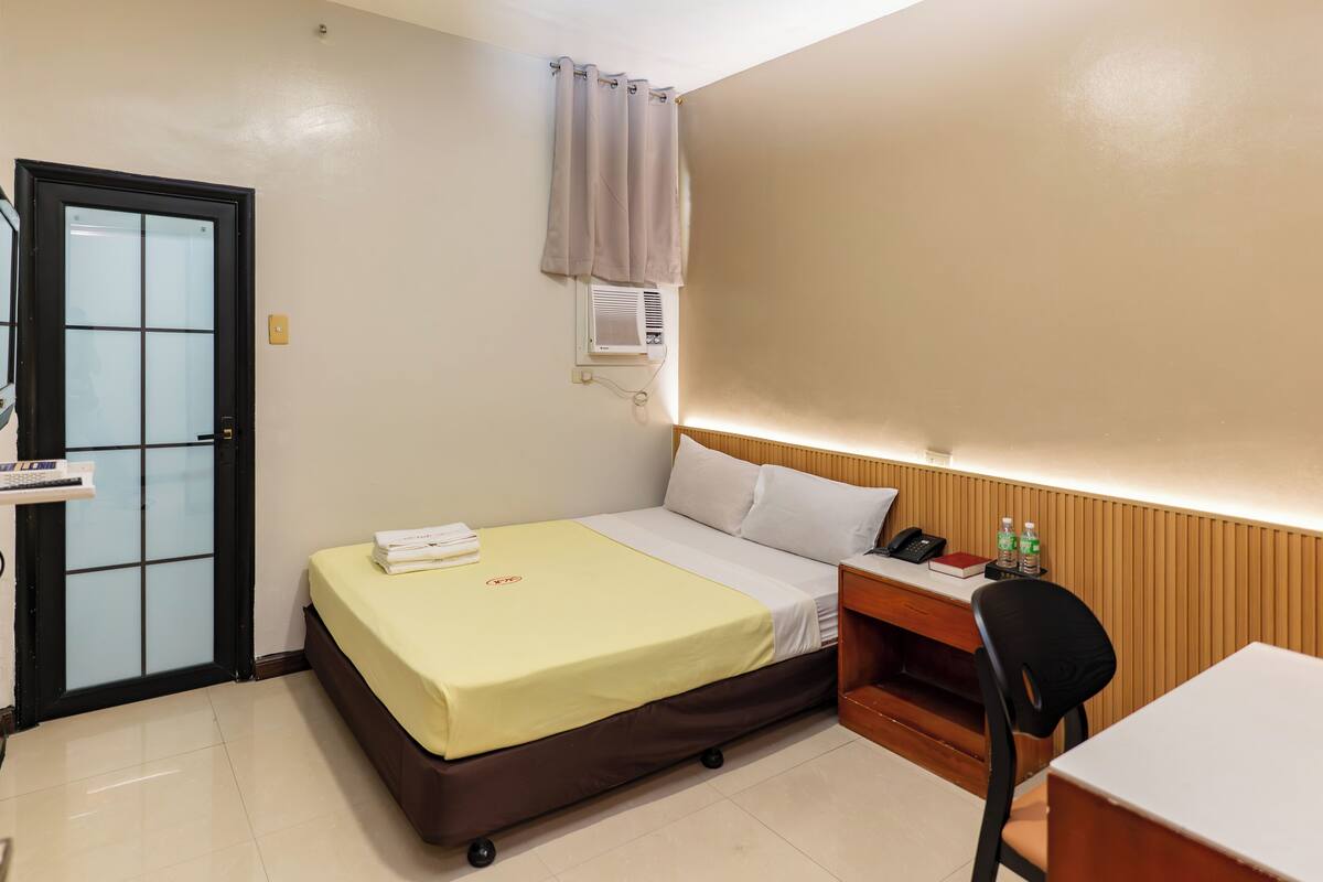 A hotel room with a bed, a desk, a chair, a door, and an air conditioning unit.