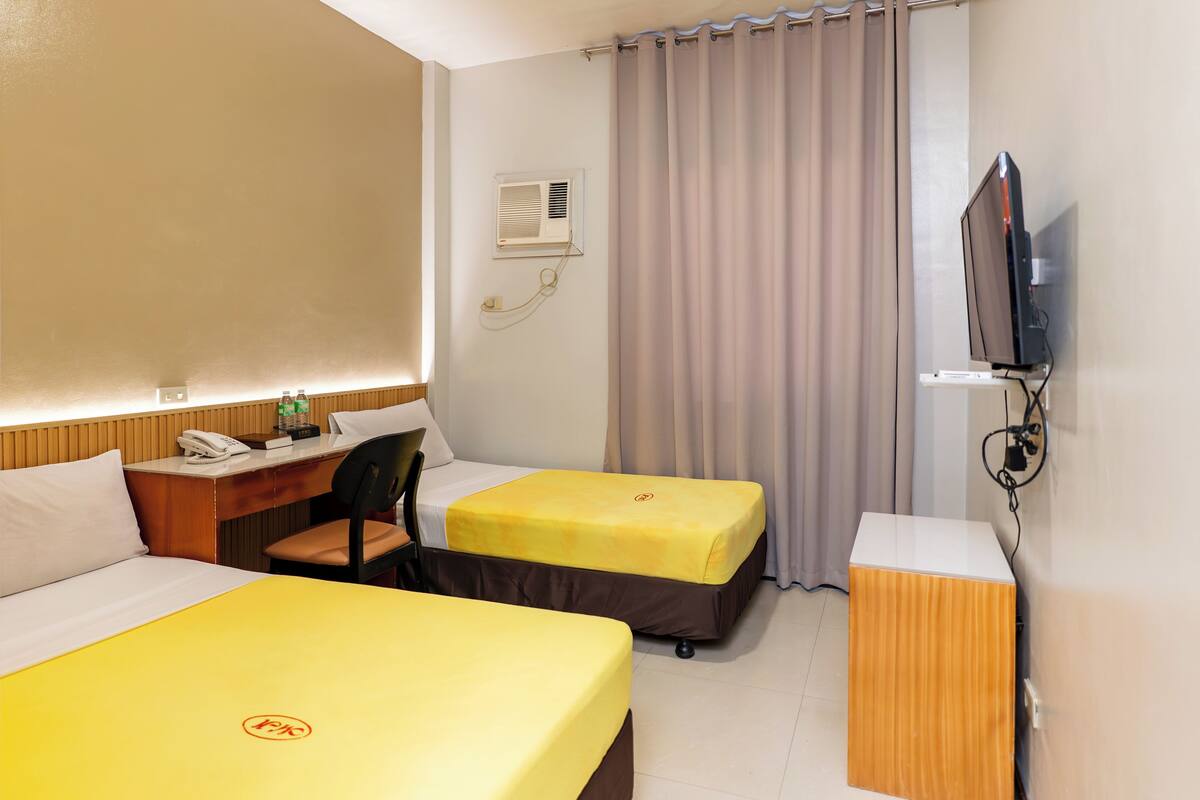 A hotel room with two beds, a desk, a chair, a TV, and an air conditioning unit.