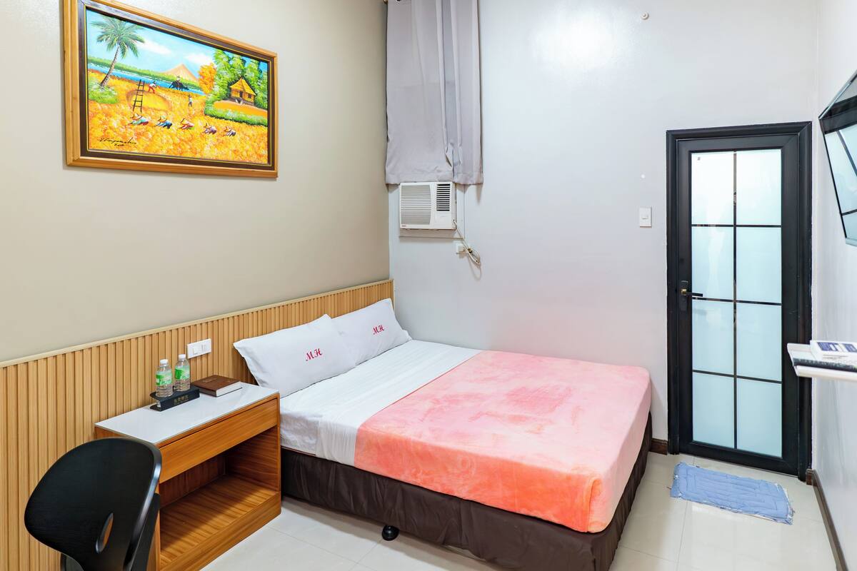 A hotel room with a bed, a desk, a chair, a painting, a door, and an air conditioning unit.