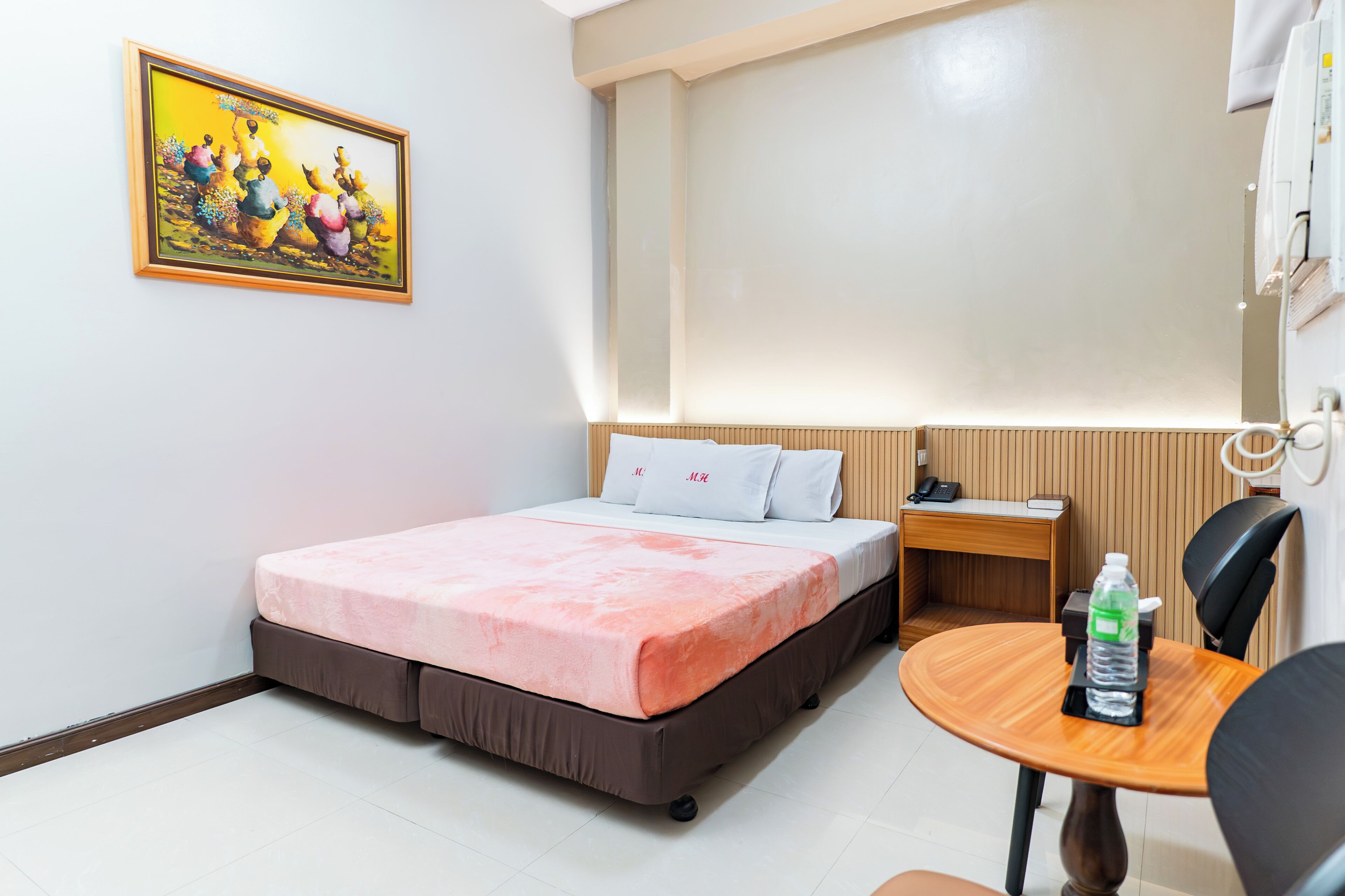 A hotel room with a bed, a small desk, a chair, a table, and a painting on the wall.