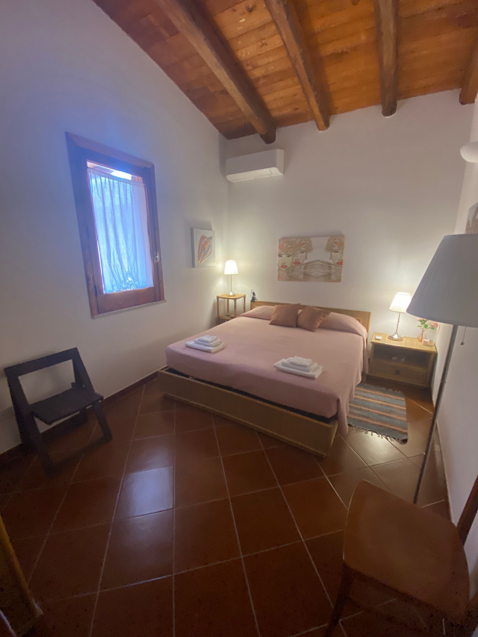 A bedroom with a bed, two bedside tables, a chair, a window with curtains, and a ceiling with wooden beams.