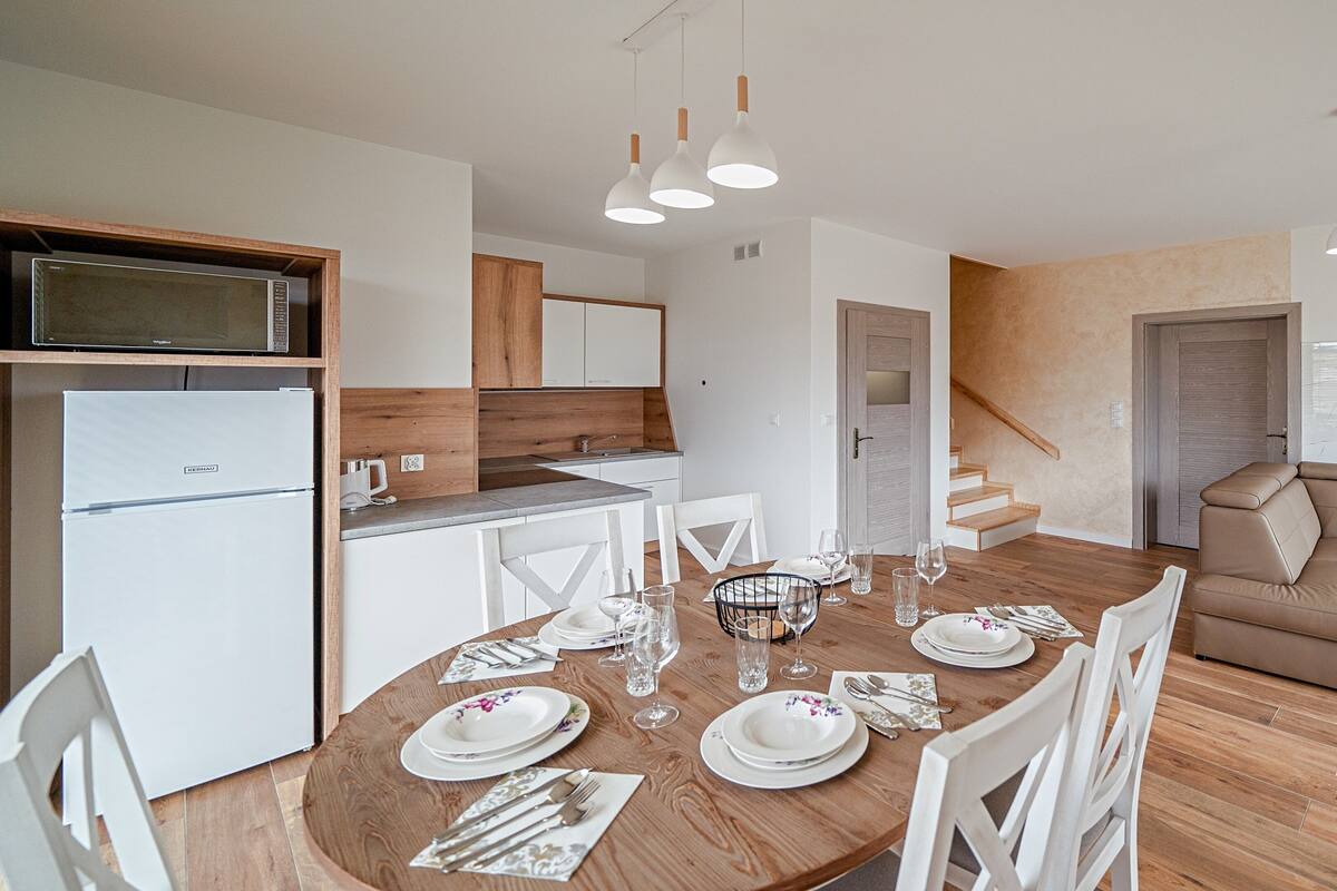 A modern kitchen with white cabinets, a built-in microwave, and a refrigerator. There is a dining area with a wooden table set for four, white chairs, and a sofa in the background.
