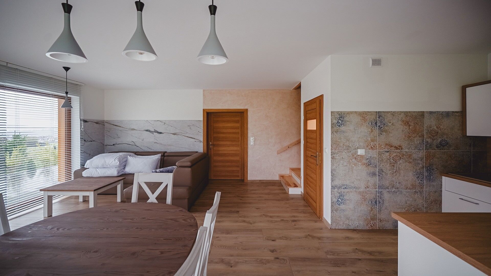 Ein modernes Schlafzimmer mit einem hölzernen Bett, einer Couch und einem Esstisch. Es gibt drei Pendelleuchten, eine Holztür und einen Küchenbereich mit gemusterten Fliesen.