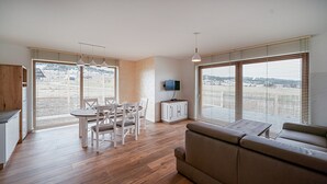 A modern living room with a sofa, dining table, and chairs. There is a TV mounted on the wall and a window with blinds. - Apartamenty Sun & Snow Kaniówka (Bukowina Tatrzanska)