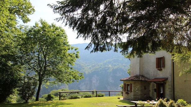 Un edificio de piedra rústico con techo de tejas rojas, rodeado de abundante vegetación y un telón de fondo de montañas panorámicas.