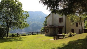 Un edificio rústico de piedra con techo inclinado, cerca de madera y un banco en una zona cubierta de césped, con montañas al fondo.
