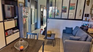 Una sala moderna con un sofá gris, una mesa de comedor de madera con una cesta de alambre, y una zona de cocina con armarios y un refrigerador.